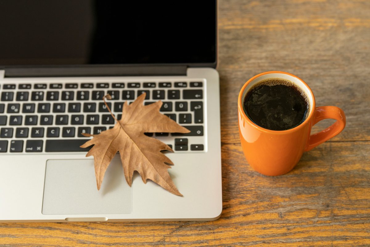 The image shows a cozy fall scene with a dry, brown maple leaf resting on a silver laptop's trackpad. Beside the laptop is an orange mug filled with black coffee, placed on a wooden surface. The warm tones of the leaf and coffee cup contrast with the sleek laptop, evoking an autumn vibe and a relaxed, work-from-home atmosphere.