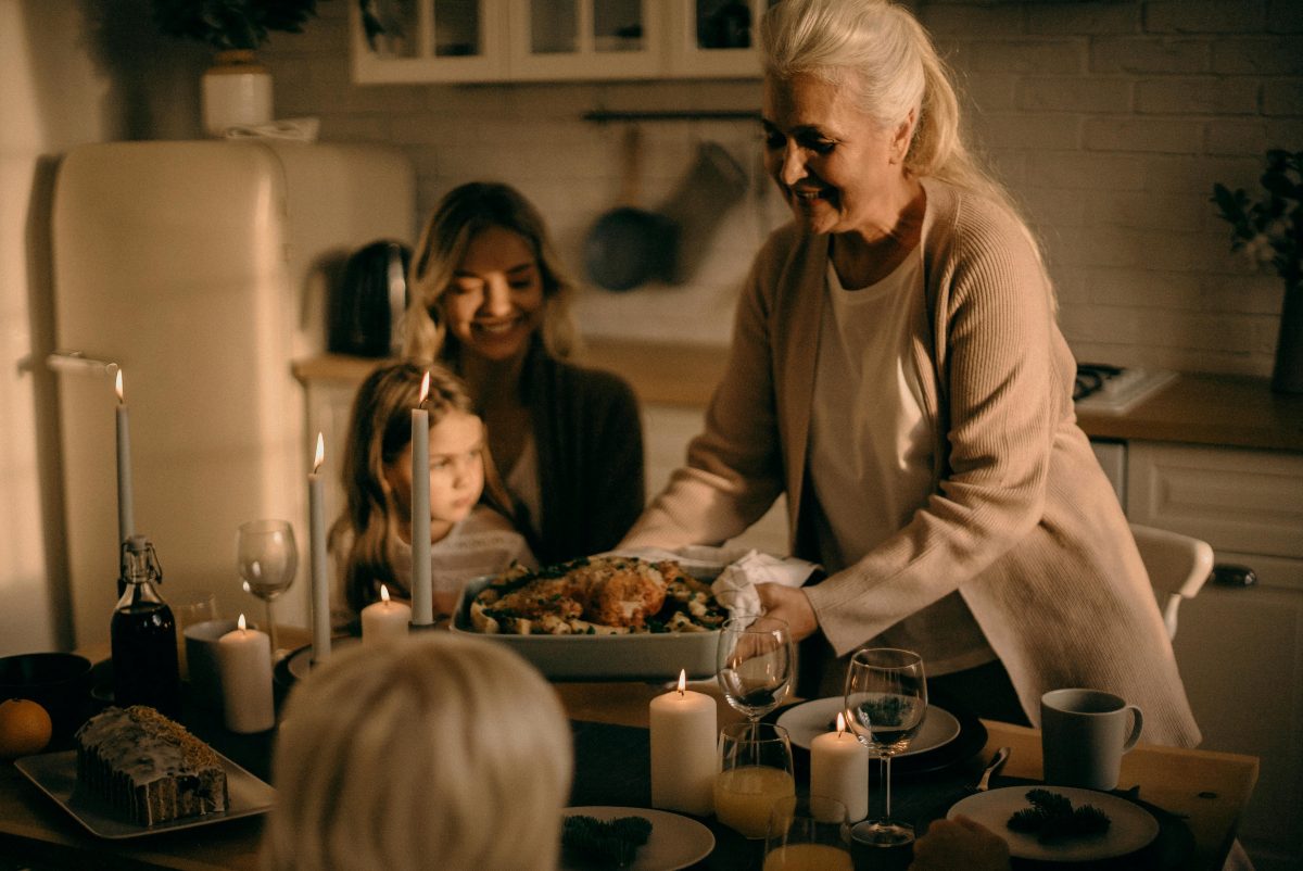 This is an image of a warm, candle-lit family gathering in a kitchen setting. A senior woman is presenting a roasted dish, surrounded by family members, including a young girl and an adult woman, who are seated at the table. The atmosphere is cozy and festive, perfect for depicting Thanksgiving or family moments of togetherness. Let me know if you'd like further help with it!