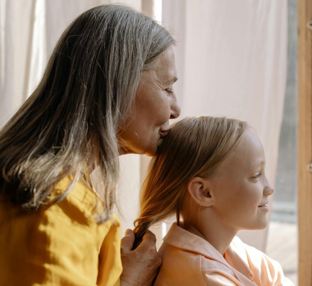 An older woman braids a young girl's hair in front of a sunlit window with white curtains, while kissing her head.