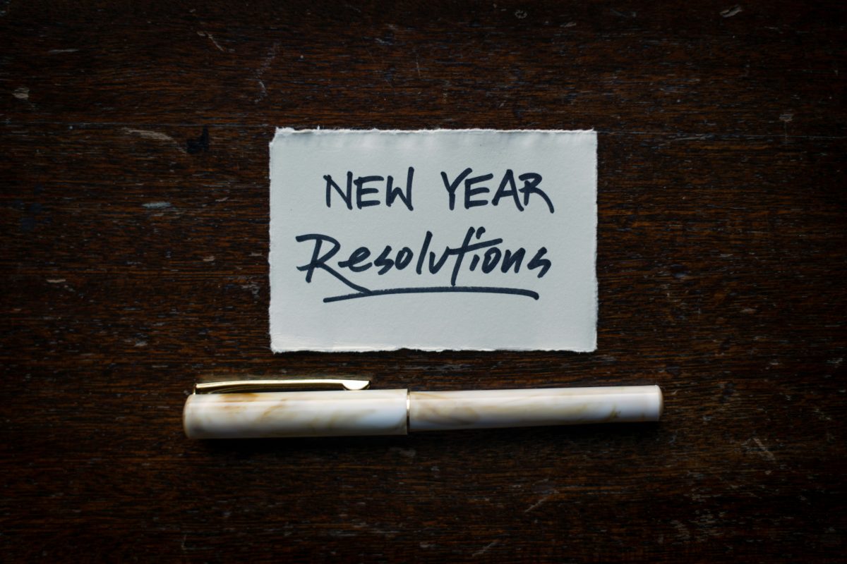This is an image with the text "New Year Resolutions" written on a piece of paper, accompanied by a pen placed below it on a wooden surface.
