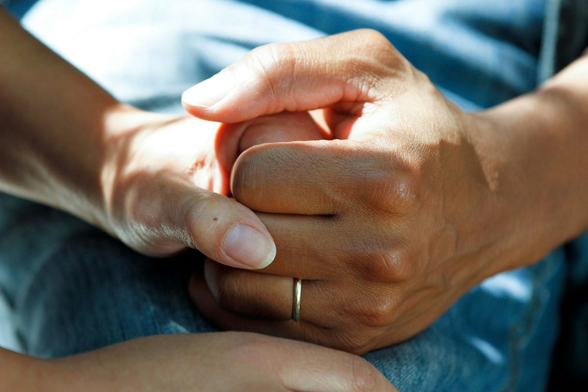 A close-up of two hands gently holding each other, conveying warmth, care, and support. One hand wears a simple gold wedding band, suggesting a connection between loved ones during a meaningful moment.