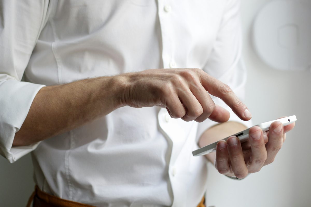 **Alt Text:** A close-up of a person wearing a white button-down shirt, using a smartphone. The individual's hands are interacting with the device, with one hand holding it and the other tapping the screen. The background is blurred, emphasizing the focus on the hands and the smartphone.