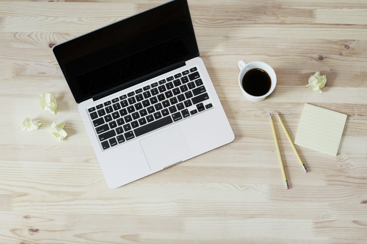 A top-down view of a workspace featuring a laptop with a black screen, a cup of black coffee, two yellow pencils, a sticky note pad, and crumpled pieces of paper on a light wooden desk.