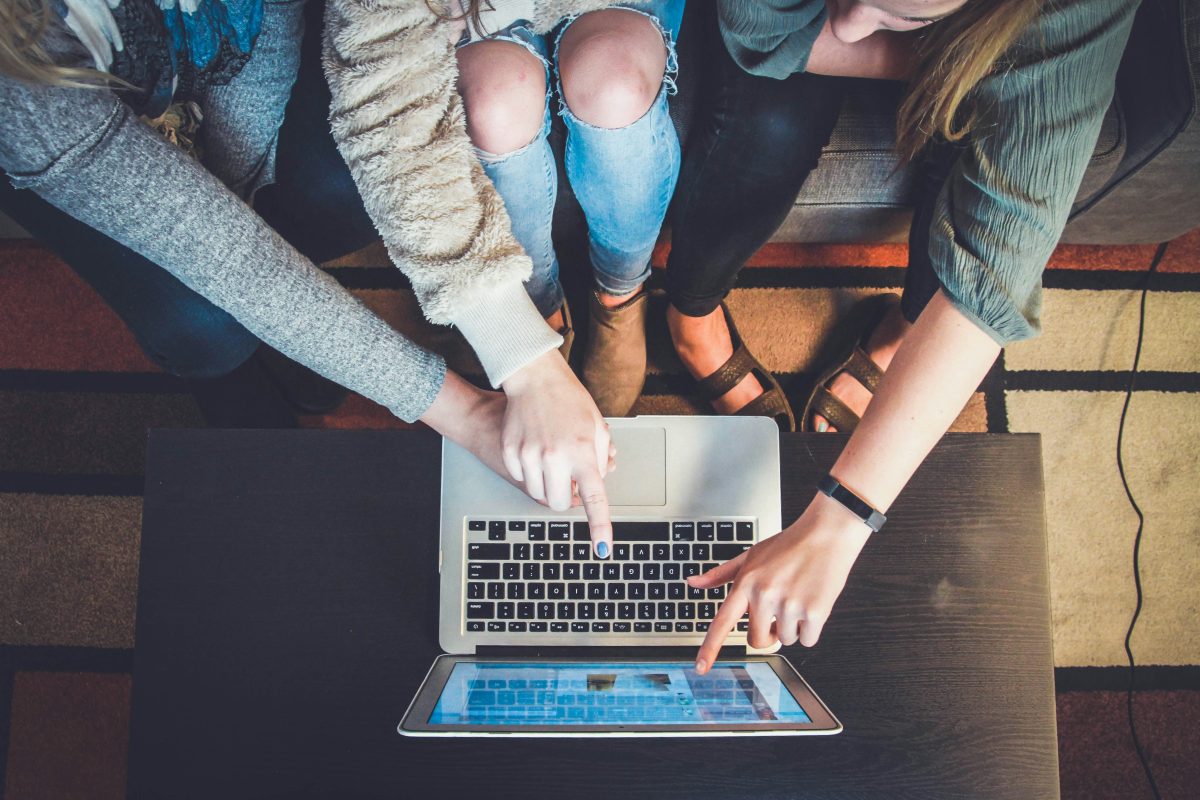 Top-down view of three people sitting around a laptop, pointing at the screen together in collaboration.