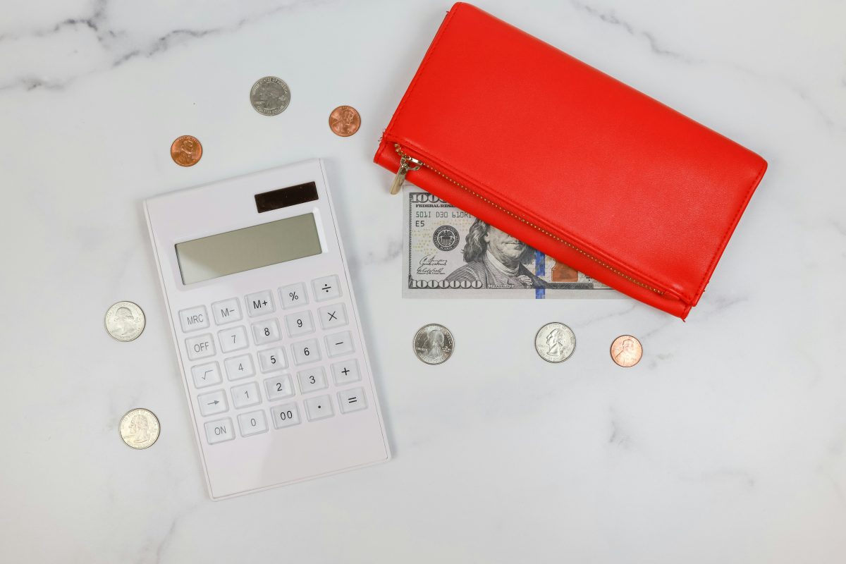 Flat lay of a white calculator, scattered U.S. coins, a visible $100 bill, and a red zippered wallet on a white marble surface.