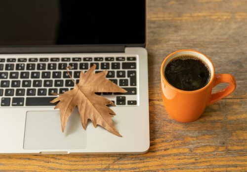 The image shows a cozy fall scene with a dry, brown maple leaf resting on a silver laptop's trackpad. Beside the laptop is an orange mug filled with black coffee, placed on a wooden surface. The warm tones of the leaf and coffee cup contrast with the sleek laptop, evoking an autumn vibe and a relaxed, work-from-home atmosphere.