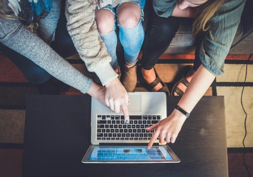 Top-down view of three people sitting around a laptop, pointing at the screen together in collaboration.