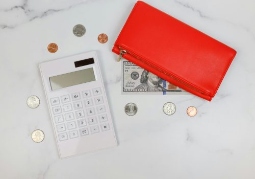 Flat lay of a white calculator, scattered U.S. coins, a visible $100 bill, and a red zippered wallet on a white marble surface.