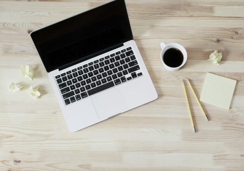 A top-down view of a workspace featuring a laptop with a black screen, a cup of black coffee, two yellow pencils, a sticky note pad, and crumpled pieces of paper on a light wooden desk.