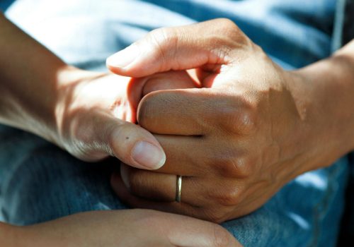 A close-up of two hands gently holding each other, conveying warmth, care, and support. One hand wears a simple gold wedding band, suggesting a connection between loved ones during a meaningful moment.