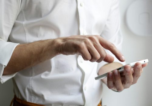 **Alt Text:** A close-up of a person wearing a white button-down shirt, using a smartphone. The individual's hands are interacting with the device, with one hand holding it and the other tapping the screen. The background is blurred, emphasizing the focus on the hands and the smartphone.