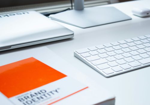 A minimalist workspace featuring a white keyboard, a closed laptop, and a monitor stand on a clean white desk. In the foreground, there is a book titled 'Brand Identity' with an orange cover, emphasizing the focus on branding.