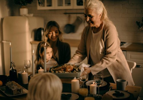 This is an image of a warm, candle-lit family gathering in a kitchen setting. A senior woman is presenting a roasted dish, surrounded by family members, including a young girl and an adult woman, who are seated at the table. The atmosphere is cozy and festive, perfect for depicting Thanksgiving or family moments of togetherness. Let me know if you'd like further help with it!
