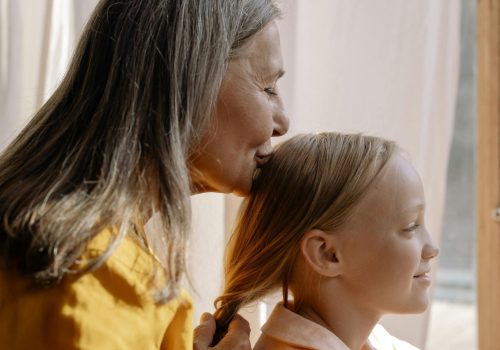 An older woman braids a young girl's hair in front of a sunlit window with white curtains, while kissing her head.