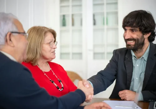 An older couple meeting with a younger financial advisor or estate planner. The advisor, a bearded man in a dark jacket, is shaking hands with an older man in glasses. An older woman in a red top sits between them, looking on. They are seated at a table in an office setting with a white cabinet visible in the background.