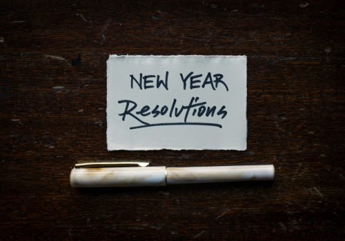 This is an image with the text "New Year Resolutions" written on a piece of paper, accompanied by a pen placed below it on a wooden surface.
